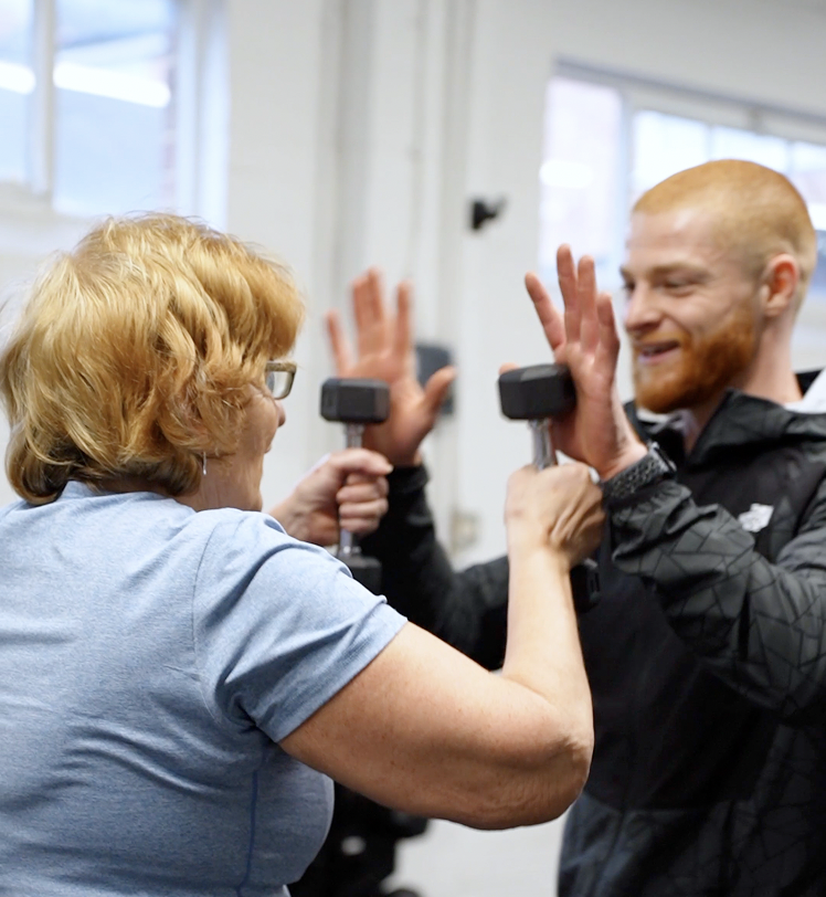 Female gym member doing dumbbell exercises with a smiling personal trainer at The Training Gyms in Bedford, highlighting supportive personal training.