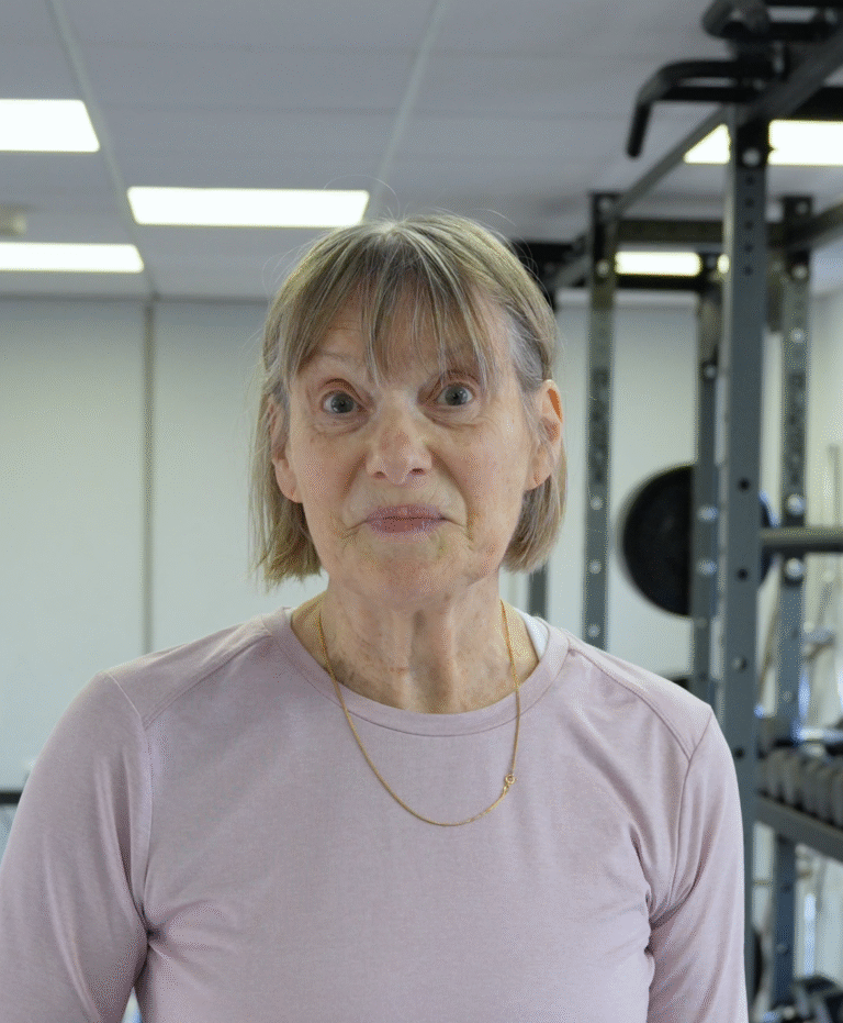 Hillary, a gym member at The Training Gyms in St Albans, smiling and standing confidently in the gym, wearing a light pink top with weightlifting equipment in the background.