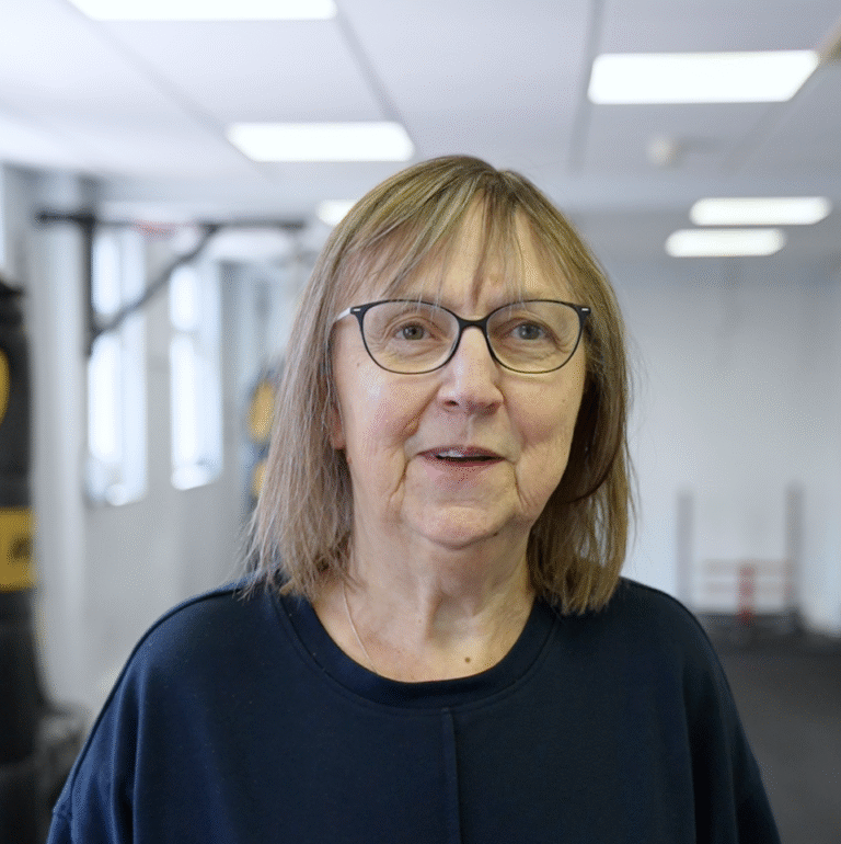 Mary Jean, wearing glasses and a navy top, standing confidently in the gym at The Training Gyms in St Albans with workout equipment behind her.