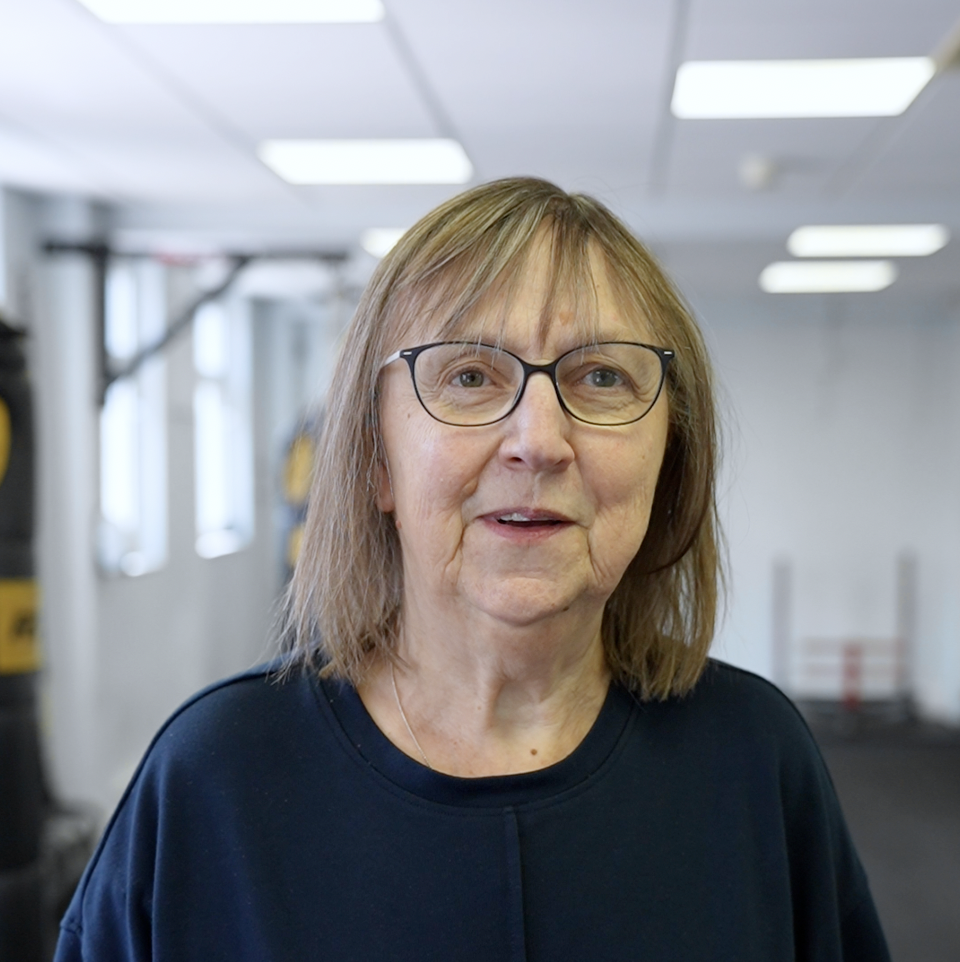 Mary Jean, wearing glasses and a navy top, standing confidently in the gym at The Training Gyms in St Albans with workout equipment behind her.