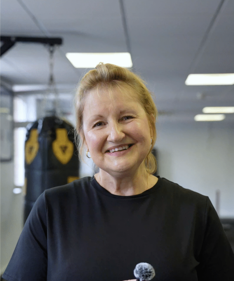 Angela smiling warmly in the gym, holding a microphone, with boxing bags in the background at The Training Gyms in St Albans.